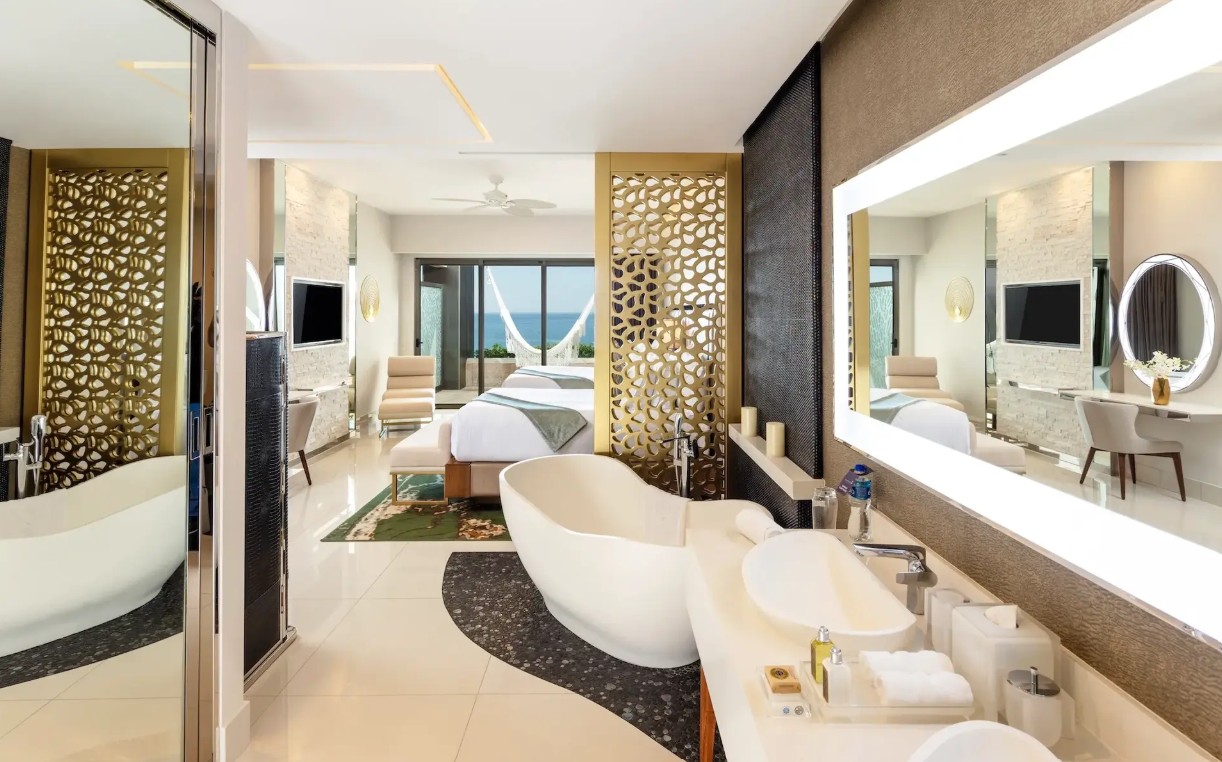 screenshot 20260728 092437 be all inclusive Luxurious hotel suite with a freestanding bathtub in the foreground, a double sink vanity with backlit mirror, and an ocean-view bedroom beyond a decorative divider.