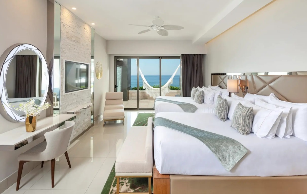 screenshot 20260728 092503 be all inclusive Spacious beachfront hotel suite with two large white beds, teal runners, and a balcony view of the ocean and a hammock outside the glass doors.