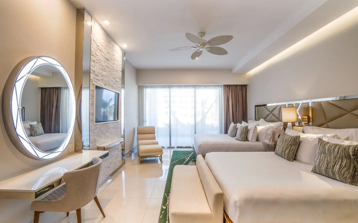 screenshot 20260728 092826 be all inclusive Two queen beds in a stylish hotel suite with a large headboard, bedside lamps, and a balcony door covered by sheer curtains.