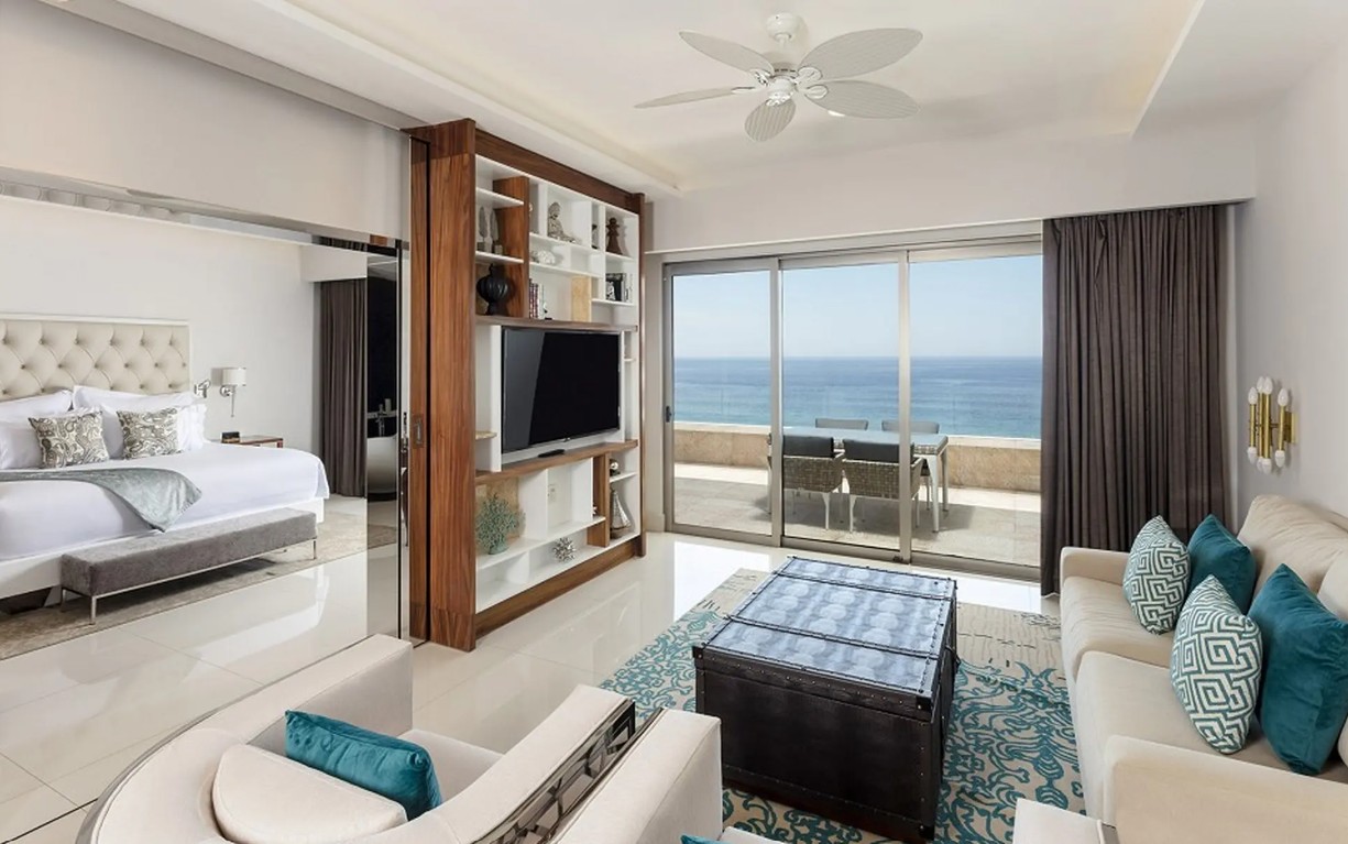 screenshot 20260728 093126 be all inclusive Luxurious ocean-view hotel suite living area with white furniture, teal cushions, and a glass door to a balcony overlooking the sea.
