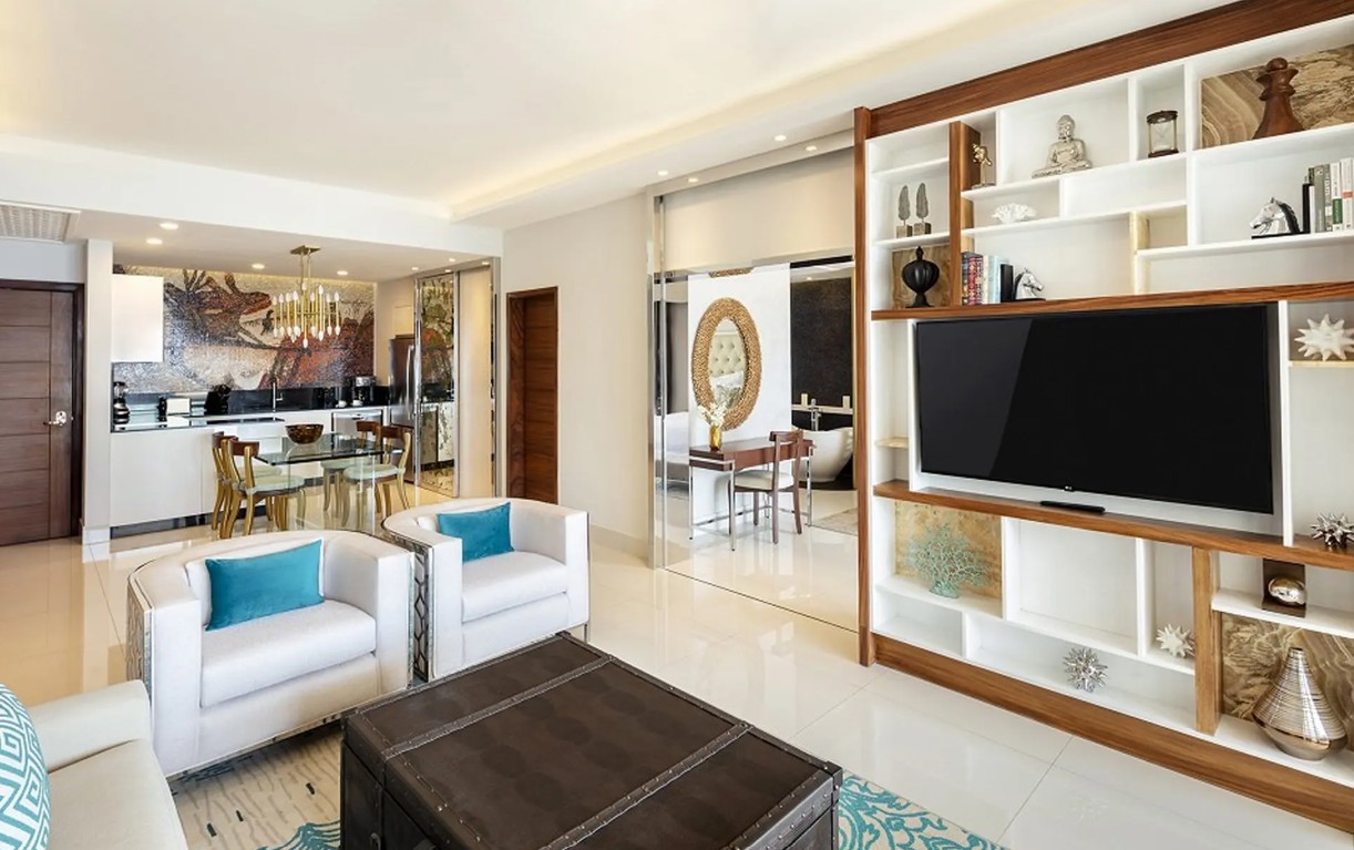 screenshot 20260728 093136 be all inclusive Open-plan living room with white sofas, teal cushions, a dark trunk coffee table, and a built-in TV wall with decorative shelving on wood trim.