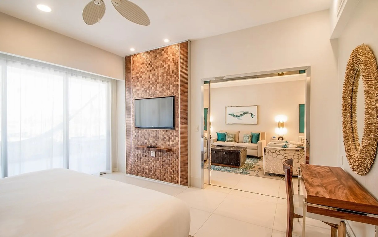 screenshot 20260728 093150 be all inclusive Hotel suite bedroom with white bedding and a mosaic TV wall, opening to a living area with teal cushions and a coffee table.