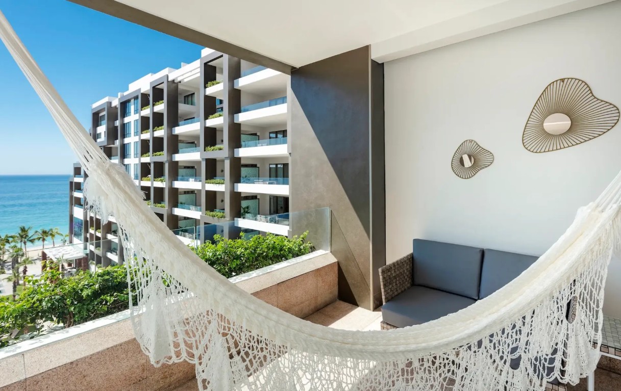 screenshot 20260728 093532 be all inclusive White hammock on a balcony with a blue ocean view and a modern hotel building nearby navigate the horizon.