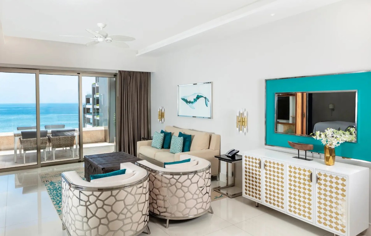 screenshot 20260728 093605 be all inclusive Bright hotel living room with white furniture, teal accents, and a turquoise-framed mirror, opening to a balcony with an ocean view