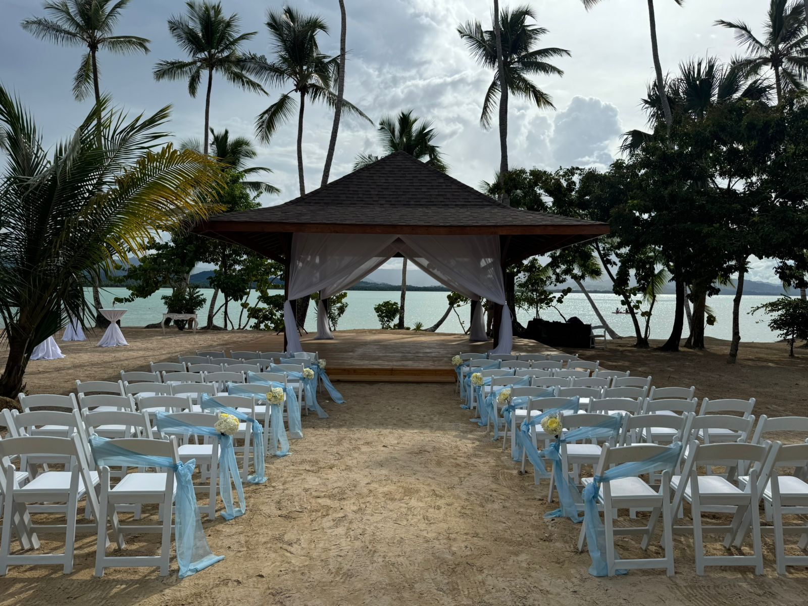 c53e8a6b796541d0844ac136336f52fd be all inclusive Beach wedding setup with rows of white chairs decorated with light blue ribbons facing a wooden pavilion on a sandy shore, ocean in the background.