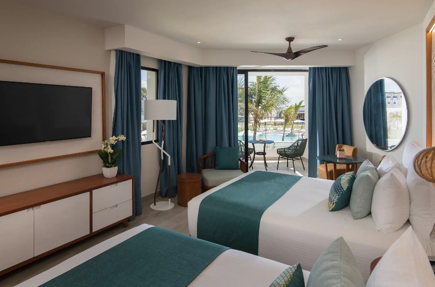 screenshot 20260805 092333 be all inclusive Hotel room with two neatly made beds and teal accents, open balcony doors showing a pool area outside