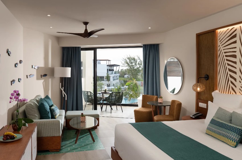 screenshot 20260805 092345 be all inclusive Hotel suite with king bed and teal accents, seating area, and balcony view through glass doors to outdoor seating area.