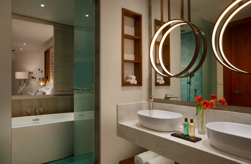 screenshot 20260805 092355 be all inclusive Hotel bathroom with double vessel sinks, circular pendant lights, and a glass-walled bathtub area to the left.