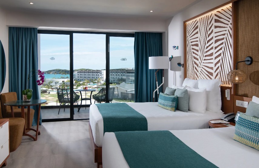 screenshot 20260805 093127 be all inclusive Hotel room with two twin beds, teal accents, and a balcony overlooking a pool and the ocean.