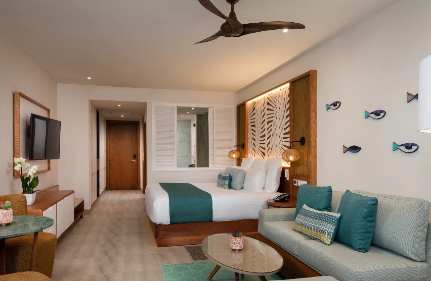 screenshot 20260805 093305 be all inclusive Hotel room with a king bed, teal accents, and a sofa; round coffee table and wall-mounted fish decorations on the right side.
