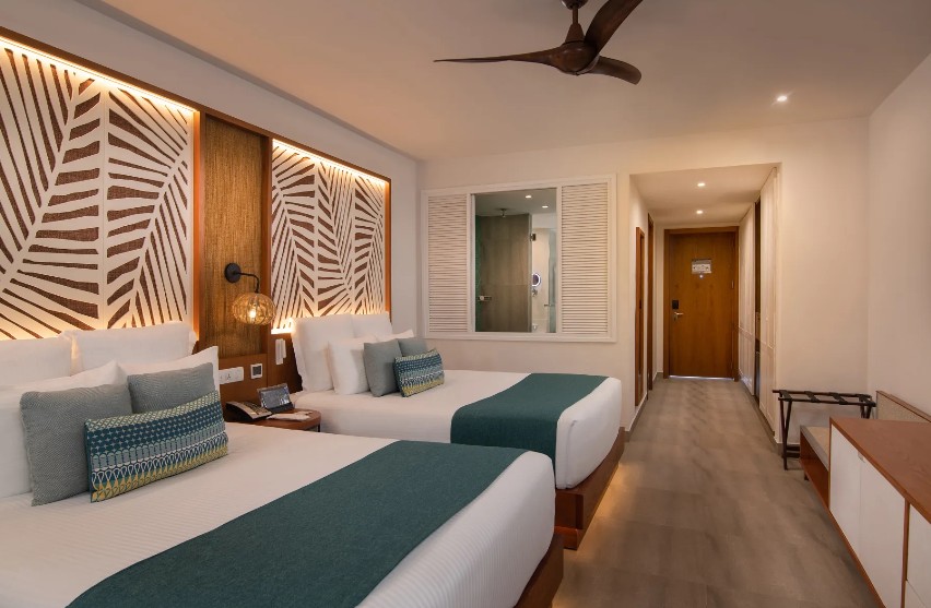 screenshot 20260805 093344 be all inclusive Two double beds in a modern hotel room with teal runners and a decorative leaf-pattern headboard between them.