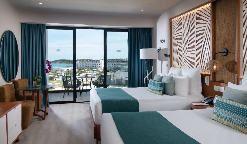 screenshot 20260805 093539 be all inclusive Hotel room with two twin beds, teal accents, and a balcony view of the ocean and resort. (informative)