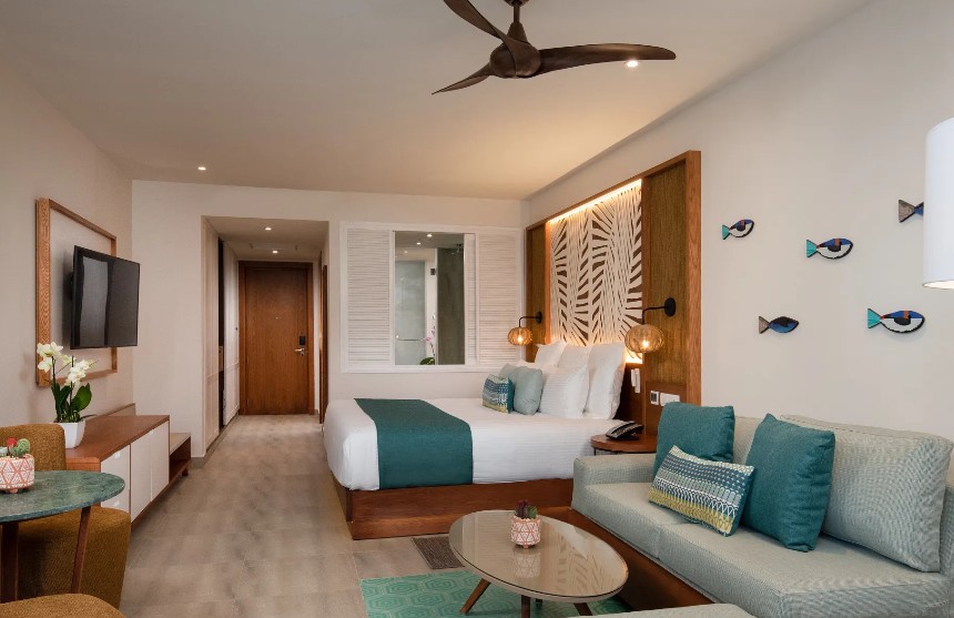 screenshot 20260805 093621 be all inclusive Hotel room with king bed, teal accents, and a seating area featuring a sofa and coffee table in a bright, modern suite