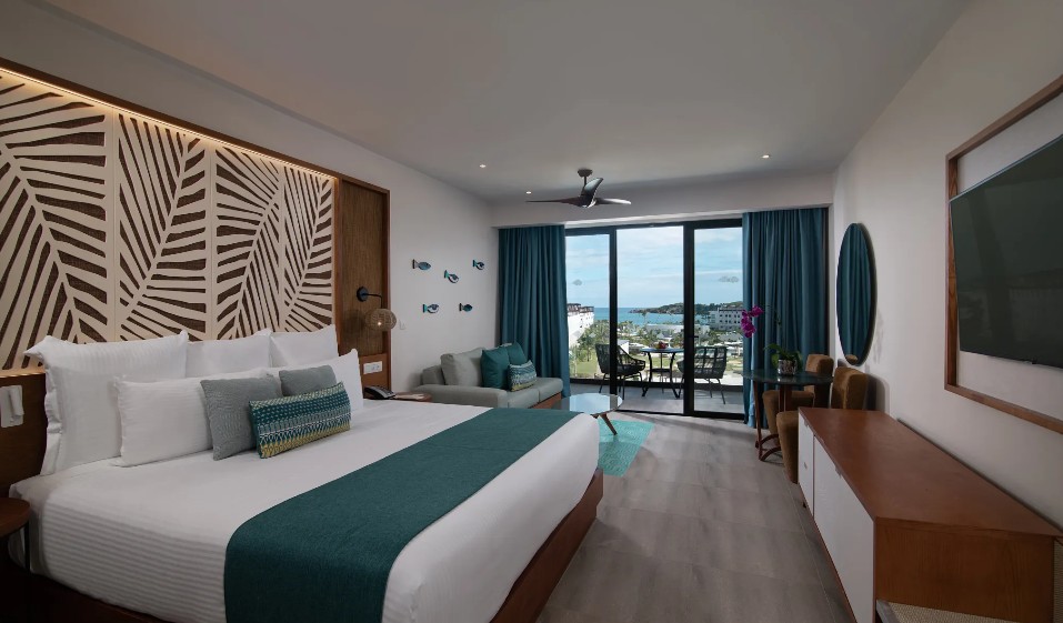 screenshot 20260805 093724 be all inclusive Hotel bedroom with a king bed, teal runner, and decorative headboard; seating area, TV, and balcony with ocean view through large glass doors.