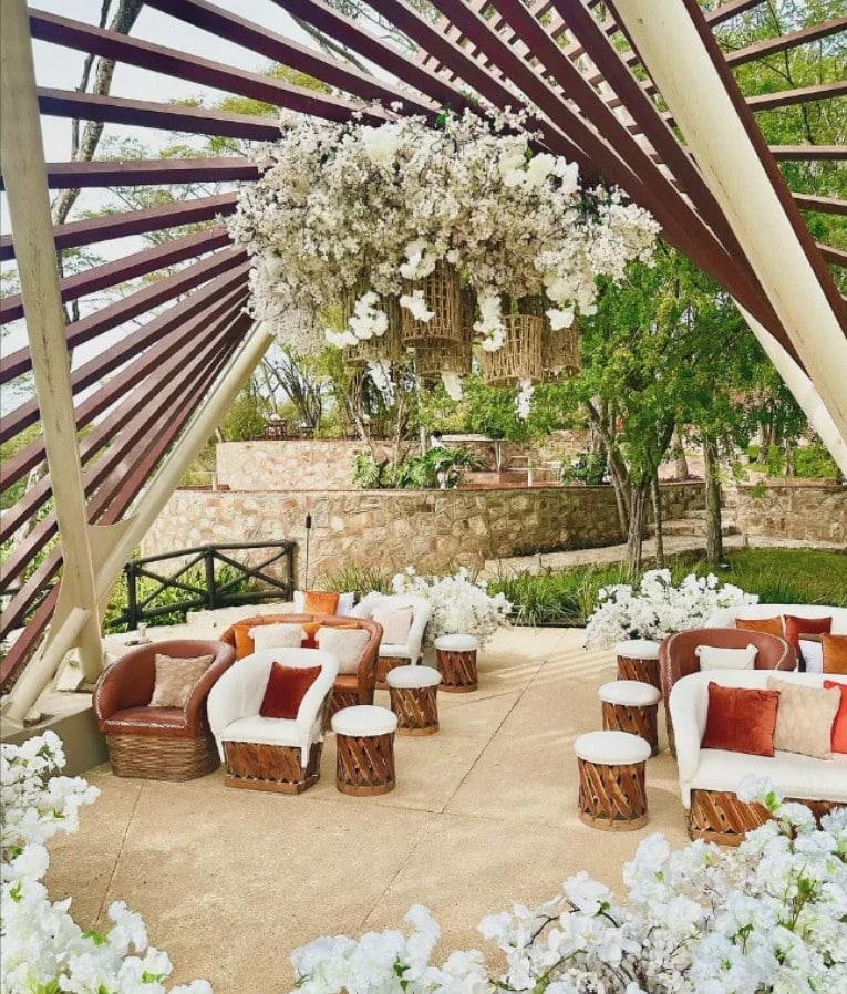 screenshot 20260819 092932 be all inclusive Outdoor lounge area under a slatted wooden pergola with white floral arrangements and wicker furniture, tan cushions, and rust pillows.