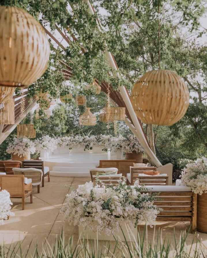 screenshot 20260819 092945 be all inclusive Outdoor ceremony space with wooden chairs, white florals, and woven lanterns hanging among trees in a garden setting.