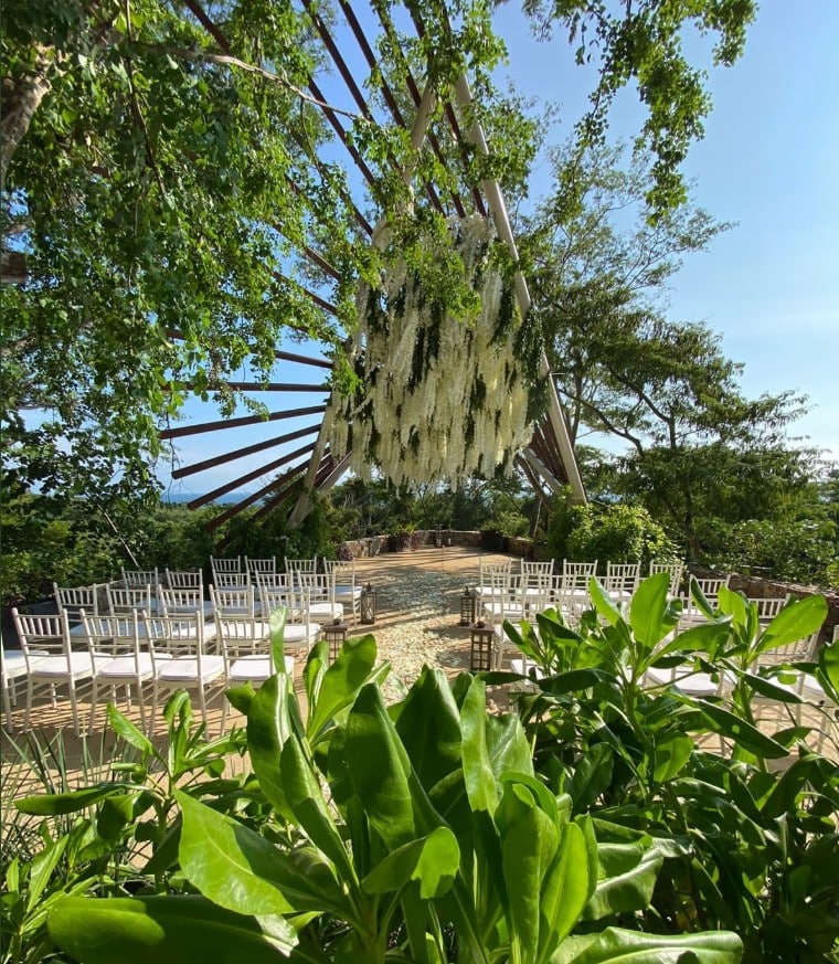 screenshot 20260819 092957 be all inclusive Outdoor wedding ceremony setup with white chairs arranged in rows beneath a wooden pergola adorned with cascading white flowers and lush greenery in bright daylight.
