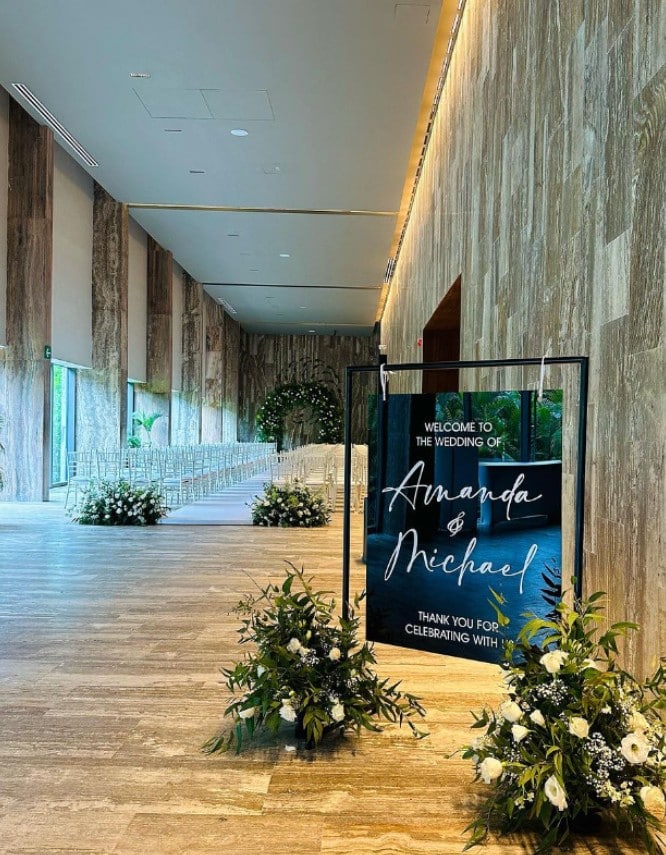 screenshot 20260819 094047 be all inclusive Wedding ceremony setup in a stone-walled hall with chairs facing an aisle and floral arrangements, plus a sign reading 'Welcome to the wedding of Amanda & Michael'