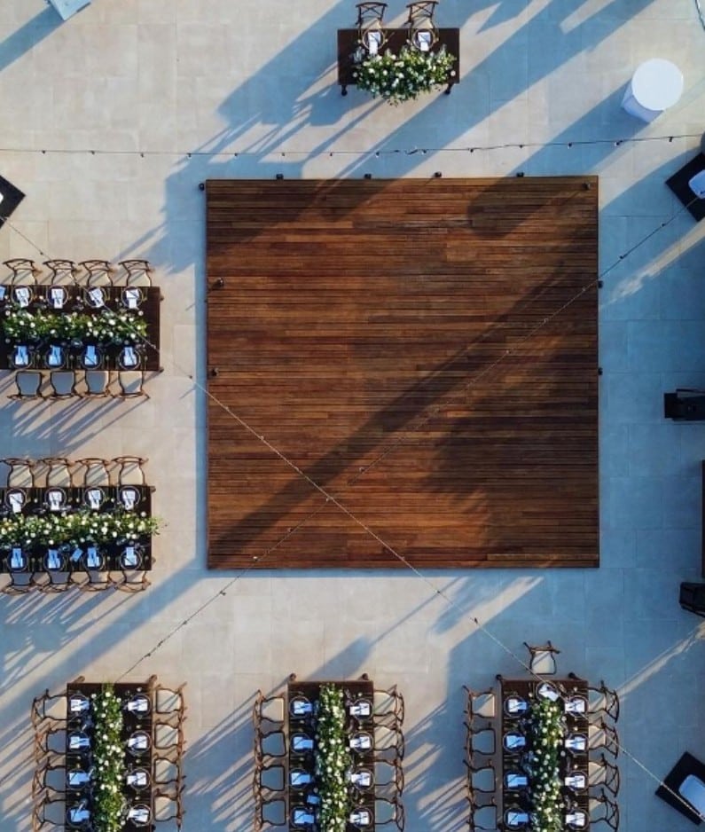 screenshot 20260819 094238 be all inclusive Aerial view of an outdoor event space with a large wooden square dance floor surrounded by long tables with chairs and flower centerpieces.