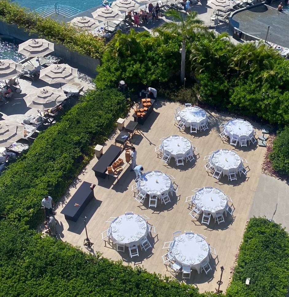 screenshot 20260819 094857 be all inclusive Aerial view of an outdoor event setup with round tables and white chairs on a wooden deck, surrounded by hedges and a pool area.