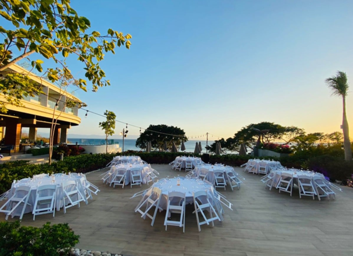 screenshot 20260819 094912 be all inclusive Outdoor seaside event setup: round tables with white chairs on a wooden deck, overlooking the ocean at sunset with string lights overhead.