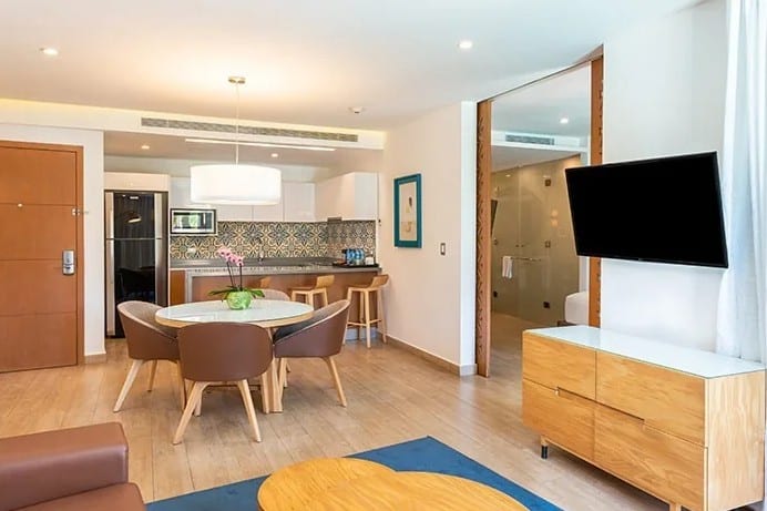 screenshot 20260928 101802 be all inclusive Open-plan living area with a round dining table and four brown chairs, adjacent kitchen counters and appliances in the background.