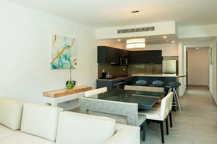 screenshot 20260928 102126 be all inclusive Open-concept living area with a white sectional, glass dining table, and a modern kitchen with dark cabinets and stainless appliances on the back wall.