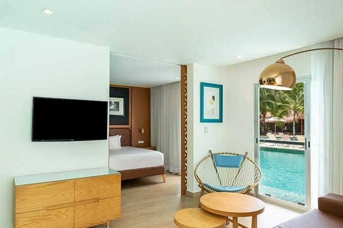 screenshot 20260928 102223 be all inclusive Bright hotel suite living area with a wall-mounted TV, wooden dresser, round coffee tables, wicker chair, and glass doors to the pool area outside.