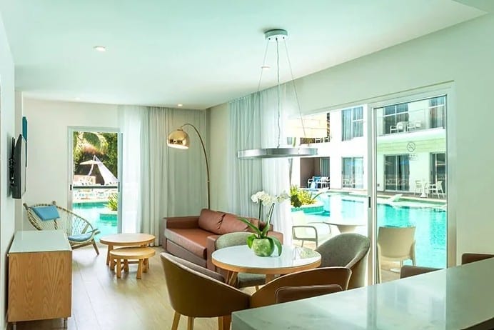 screenshot 20260928 102245 be all inclusive Bright modern living room with large windows overlooking a pool, light wood furniture, brown sofa, and round coffee tables.