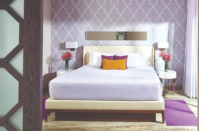 screenshot 20260928 102532 be all inclusive Modern hotel-style bedroom with a large bed, white bedding, purple geometric wallpaper, and a purple rug.