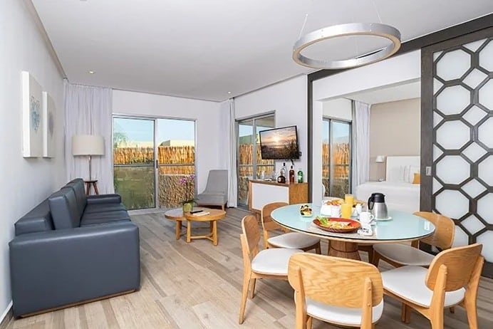 screenshot 20260928 102541 be all inclusive Bright open-plan living and dining room with a round glass table, six wooden chairs, a gray sofa, and large windows to the yard.