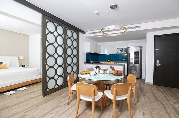 screenshot 20260928 102551 be all inclusive Open-plan hotel suite with a round glass dining table for six, wooden chairs, a compact kitchen with a blue backsplash, and a bedroom partially visible beyond a geometric divider.