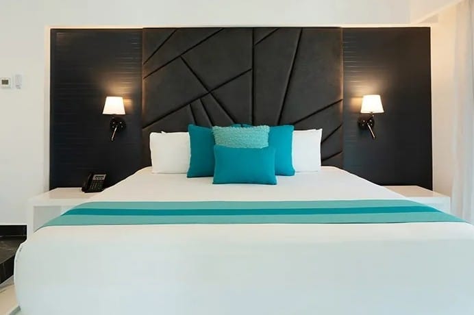 screenshot 20260928 102730 be all inclusive King bed with white sheets and teal throw pillows in a modern hotel room, two wall lamps and a dark geometric headboard.