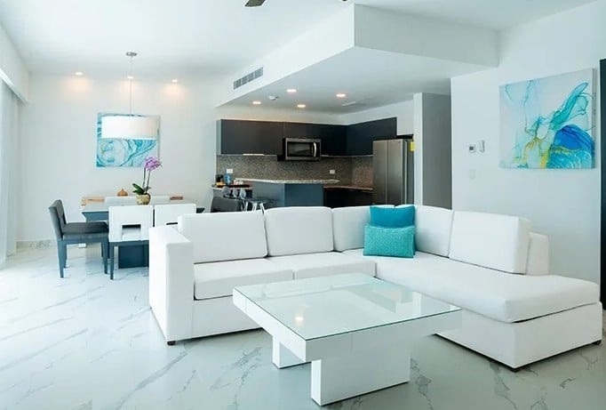 screenshot 20260928 102739 be all inclusive Open-concept living area with a white sectional sofa, glass coffee table, and turquoise accent pillow; kitchen in back.