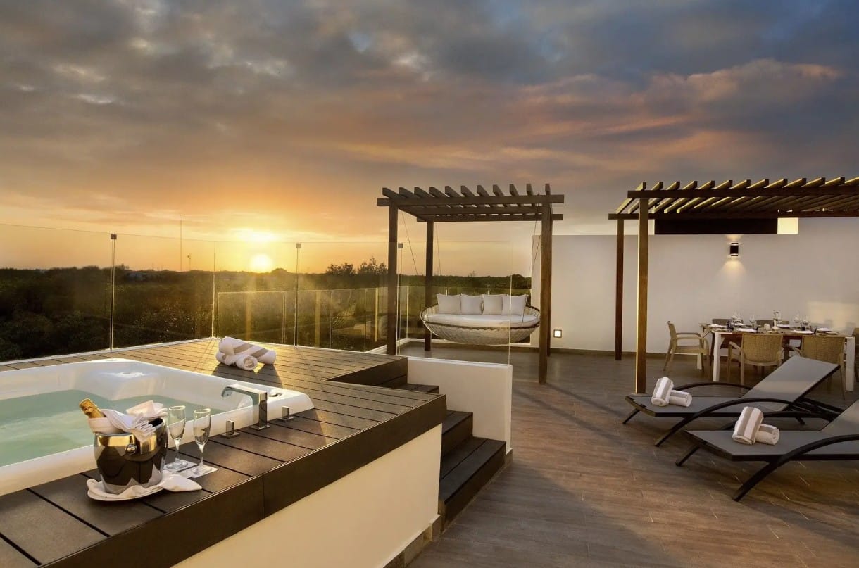 screenshot 20260928 102751 be all inclusive Rooftop terrace at sunset with a hot tub, glass railing, lounge chairs, a hanging swing, and a dining area under wooden pergolas.