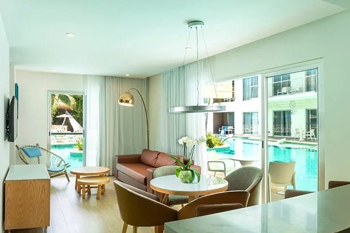 screenshot 20260928 102928 be all inclusive Bright open living room with a brown sofa, round glass table, and seating area; large windows overlook a pool outside.