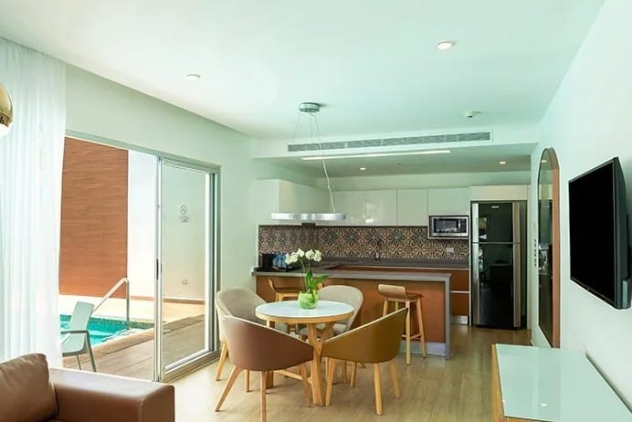 screenshot 20260928 102937 be all inclusive Open-plan living area with a round dining table and four chairs near a kitchen island, sliding glass door to a backyard pool on the left, and a wall-mounted TV on the right; bright, modern decor with a patterned backsplash and greenish lighting.