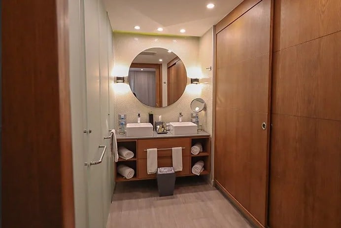 screenshot 20260928 102947 be all inclusive Hotel bathroom with double sinks, round mirror, and wooden cabinetry; towels neatly arranged on open shelves and a trash bin below the counter.