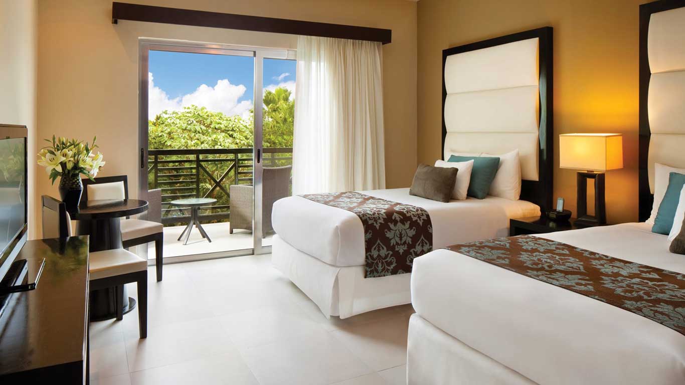 slide6 be all inclusive Hotel room with two white beds, brown patterned runners, and teal/brown throw pillows, beside a nightstand with a lamp.