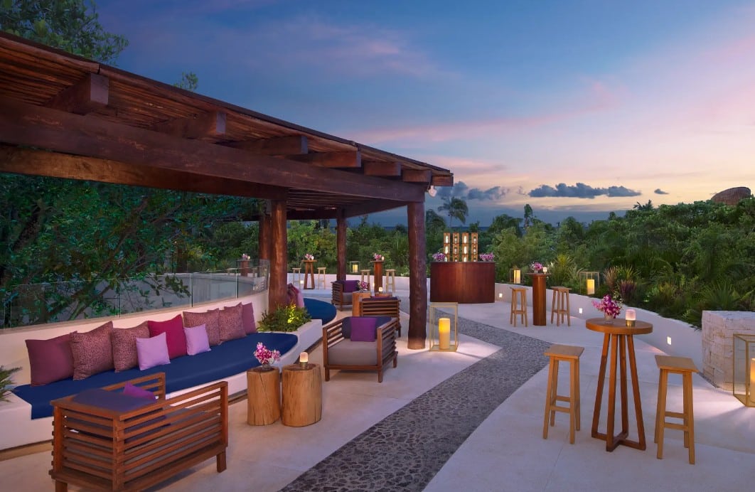 screenshot 20261007 130300 be all inclusive Outdoor lounge under a wooden pergola at dusk with purple-pink cushions and lanterns along a stone path