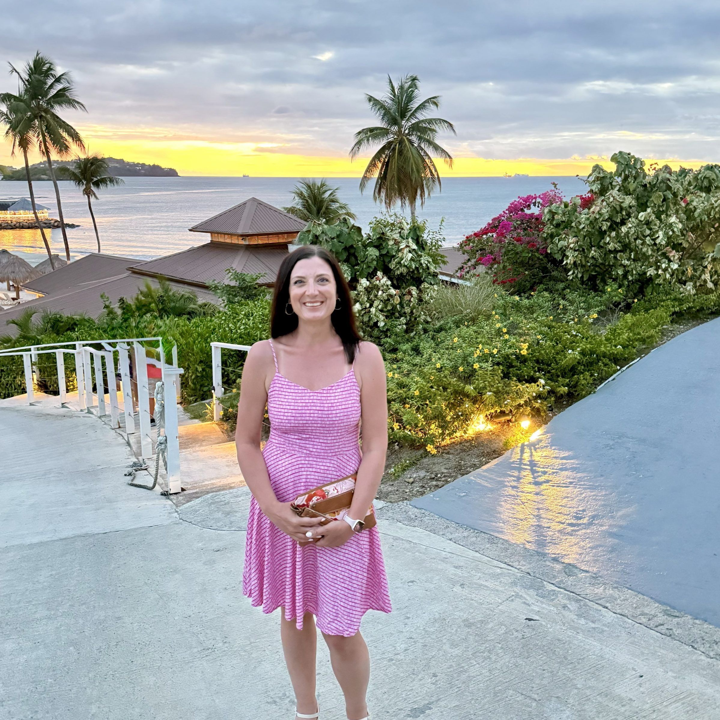 Woman in a pink striped dress standing on a seaside walkway at sunset, with palm trees and calm ocean in the background.